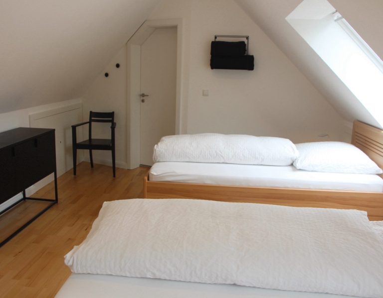 Helle Dachzimmer mit zwei Betten, einem Stuhl und einem Sideboard.