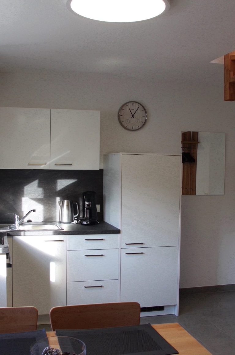 Helle Küche mit weißen Möbeln, Herd, Kühlschrank und Wanduhr.